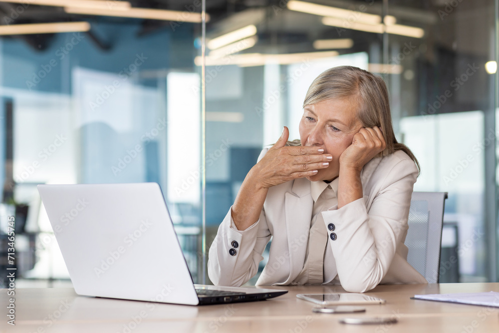Tired senior woman sitting at desk in front of laptop in office in business suit, head on hand and yawning while covering mouth with hand. Tired senior woman sitting at desk in front of laptop in office in business suit, head on hand and yawning while covering mouth with hand.