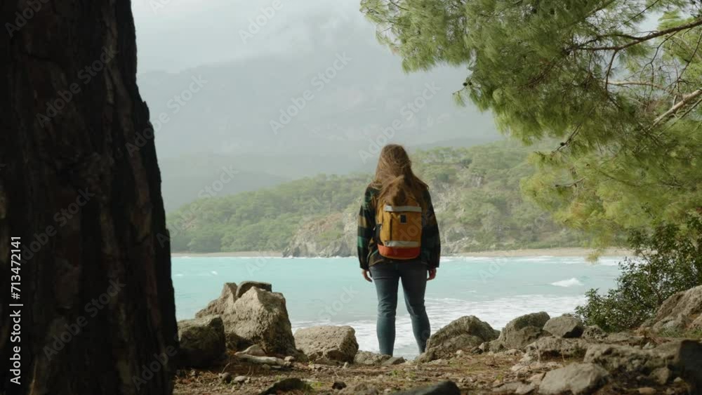The camera follows a woman with a backpack on her journey as she walks through the pines, steps on stones, and emerges into a bay with turquoise water. Windy weather. The concept of solitude and a som