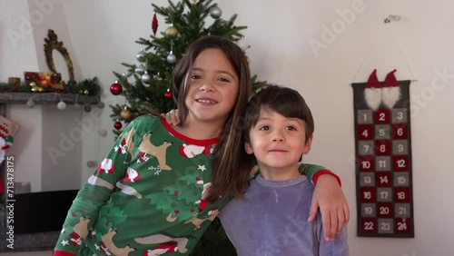 Siblings posing for camera with Christmas tree in background. sister and brother wearing pajamas smiling