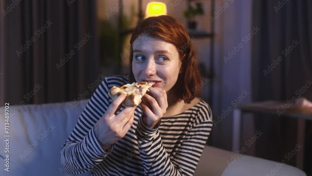Close up portrait of young red haired woman looking around to make sure nobody watching and eating pizza with great pleasure sitting on sofa at home at night Break the diet or excellent food concept Close up portrait of young red haired woman looking around to make sure nobody watching and eating pizza with great pleasure sitting on sofa at home at night Break the diet or excellent food concept