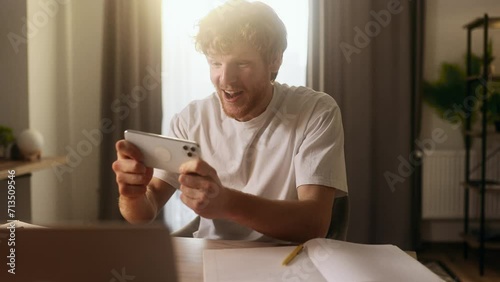 Handsome red haired young freelancer relaxing during break and playing in online video games on smartphone holding in horizontal landscape mode during work day at home Relaxation and job concept photo