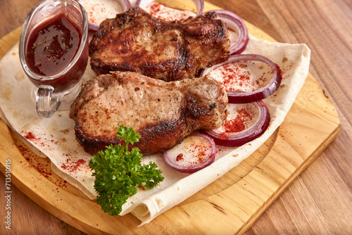 Part of the wooden board with fried beef meat with red onion rings, sauce and pita bread on the wooden table, close-up perspective view, shallow depth of field. Meat and onion in focus