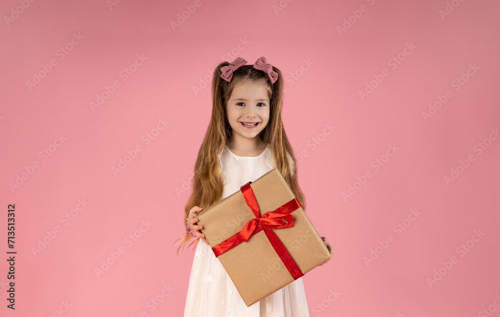 a little blonde girl with long hair holds a gift in her hands and smiles on a pink background. The girl looks out from behind the gift. The concept of a birthday party a little blonde girl with long hair holds a gift in her hands and smiles on a pink background. The girl looks out from behind the gift. The concept of a birthday party