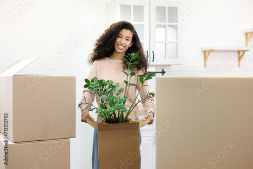 happy young African American woman is moving into new home. person stands among cardboard boxes and smiles, holding a houseplant, mortgage or rental property in his hands. mover service for moving. © MyJuly