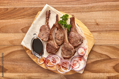 Fried lamb ribs meat with red onion rings, sauce and pita bread on the wooden board on the wooden table, flat lay top view