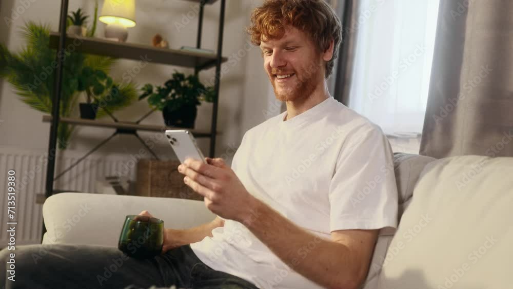 Attractive young man with cup of hot coffee or tea while hold smartphone checking email watching social media indoors Smiling red haired guy texting on phone relaxing at home Attractive young man with cup of hot coffee or tea while hold smartphone checking email watching social media indoors Smiling red haired guy texting on phone relaxing at home