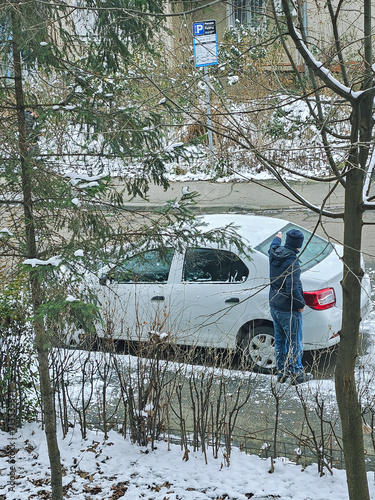 A man cleaning a car from snow on a winter day with a defrost liquid. A Caucasian man cleans his car after a snowfall on a frosty day. Removing snow from windows with a fluid solution.