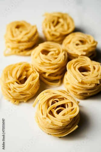 Dry tagliolini or tagliatelle pasta in nests in the bowl on white marble background. Uncooked ingredient