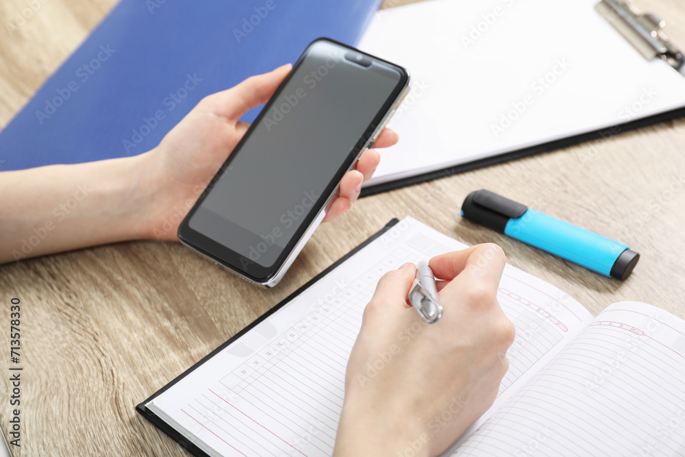 Woman taking notes while using smartphone at wooden table, closeup
