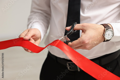 Man cutting red ribbon with scissors on blurred background, closeup photo