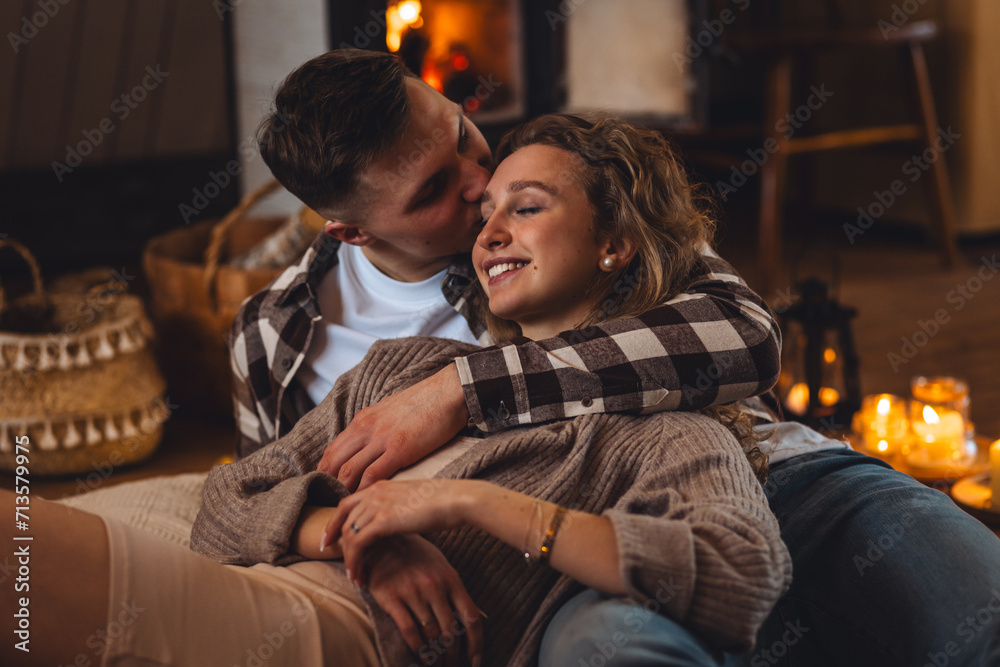 Beautiful young loving couple relaxing near fireplace in bed at home. Happy spouses enjoying lazy romantic winter cozy morning in bedroom. Candles, garlands, countryside interior. Romantic date - obrazy, fototapety, plakaty 