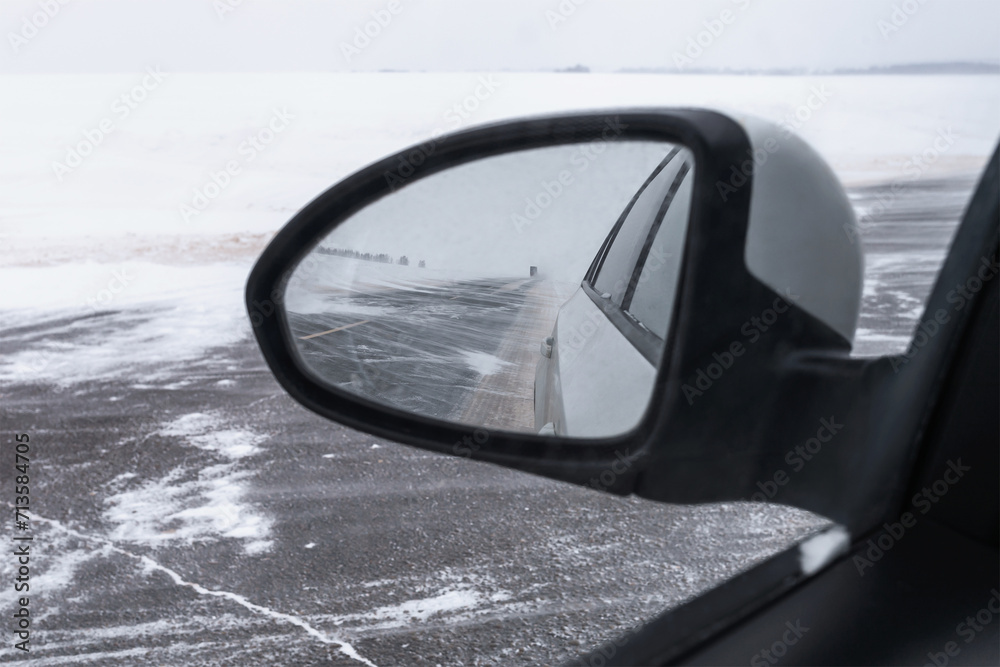 View from the rear view mirror of a car on snowfall and blizzard, the road is partially covered with snow