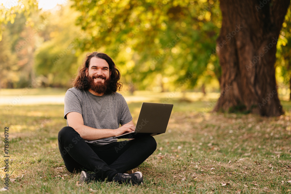 Φωτογραφία Wide smiling bearded man with long hair sits in park and works at his laptop on a sunny day