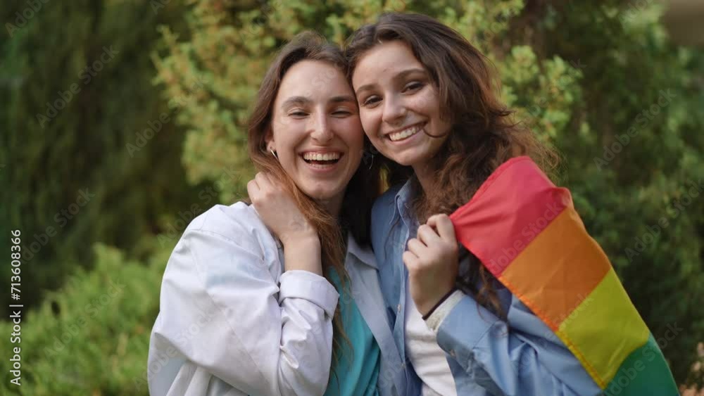 Two girls stand hugging each other in a city park. The girls wrapped themselves in a rainbow flag. Teenagers laugh and look at the camera while standing among green trees on a bright sunny summer day Two girls stand hugging each other in a city park. The girls wrapped themselves in a rainbow flag. Teenagers laugh and look at the camera while standing among green trees on a bright sunny summer day