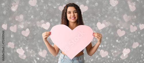Beautiful young woman with pink paper heart on grey background with glowing lights. Valentines Day celebration