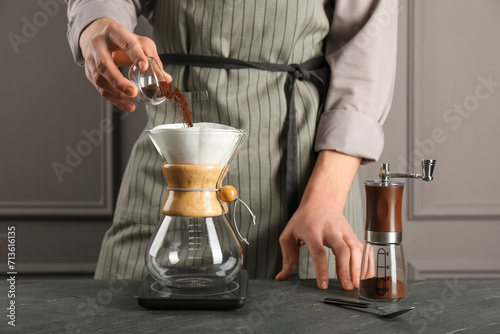 Man adding coffee into glass chemex coffeemaker with paper filter at gray table, closeup