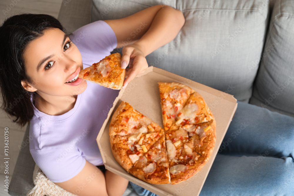 Young woman eating piece of tasty pizza and holding cardboard box on sofa at home