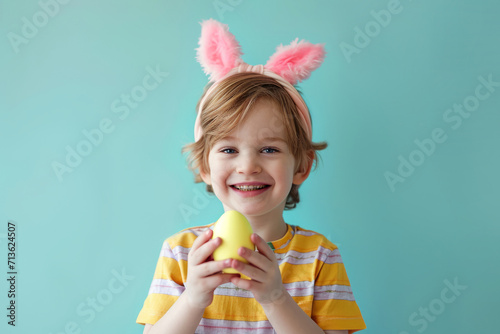 Happy small boy with bunny ears holding colored Easter egg against blue background