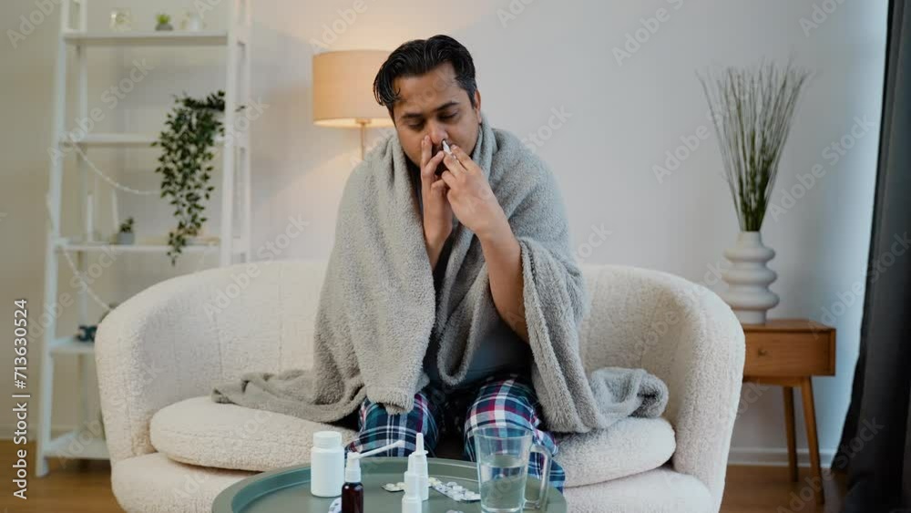Indian man wrapped in a blanket sprays a nasal spray while sitting on sofa Indian man wrapped in a blanket sprays a nasal spray while sitting on sofa
