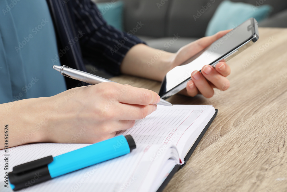 Woman taking notes while using smartphone at wooden table indoors, closeup Woman taking notes while using smartphone at wooden table indoors, closeup
