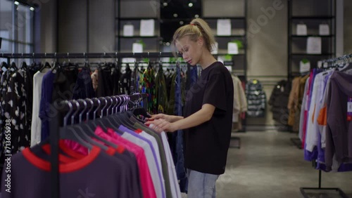 A young blonde woman with fair skin chooses a T-shirt in a clothing store, a woman shopper in a clothing store, a woman in a black T-shirt in a store is sorting through clothes on a rail.