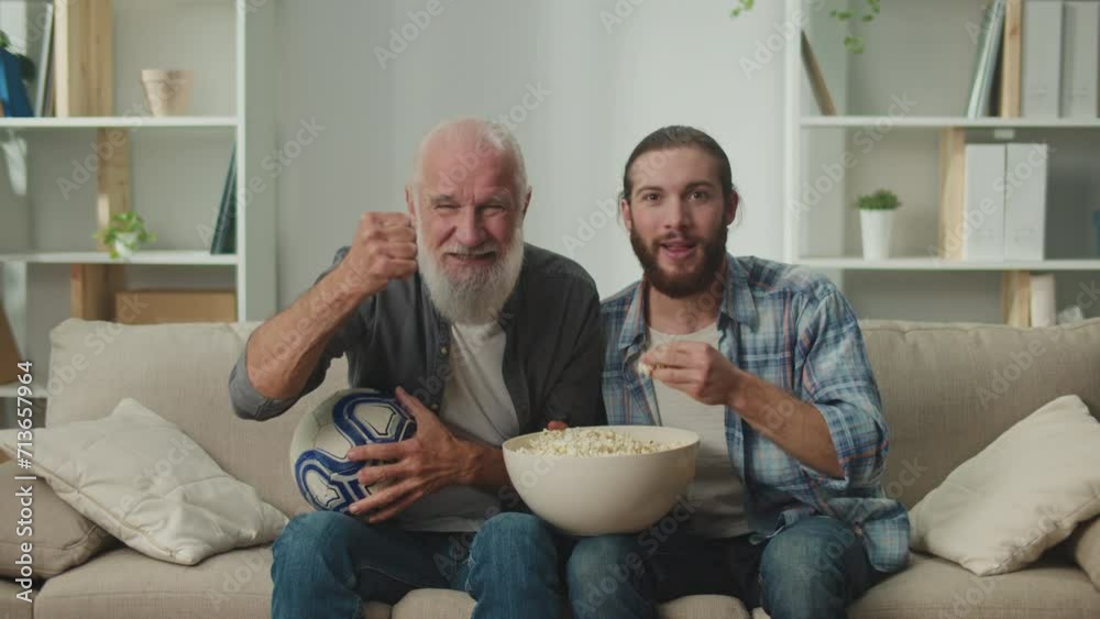 Elder and young fan enjoying a game together on a sofa, with snacks and drinks, engrossed in online match streaming, family bonding, and shared sports enthusiasm Elder and young fan enjoying a game together on a sofa, with snacks and drinks, engrossed in online match streaming, family bonding, and shared sports enthusiasm