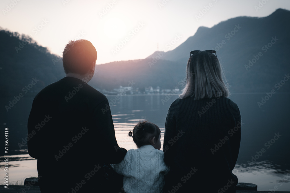 Silhouette of a family - father, mother and daughter sitting on the lake or sea beach with their backs to the camera and looking to the horizon on the sunset. Love and happy family concept.