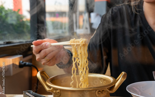 A woman's hand forks hot, smoking Korean noodles in a brass pot at a restaurant.