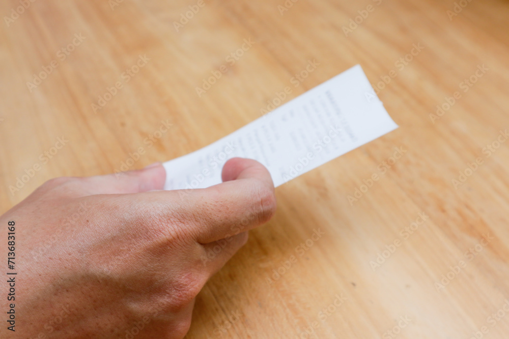 A man's hand is holding a shopping receipt on a wooden table A man's hand is holding a shopping receipt on a wooden table