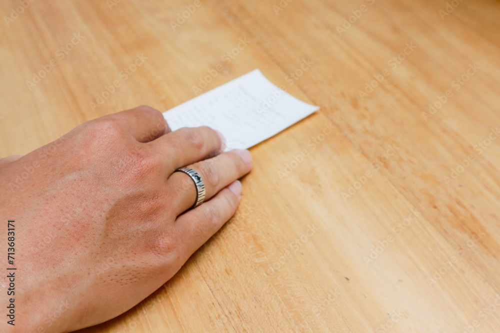 A man's hand is holding a shopping receipt on a wooden table A man's hand is holding a shopping receipt on a wooden table
