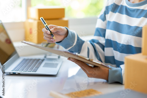 Young Asian woman starting an online shopping business at home using laptop is checking internet orders and writing notes.