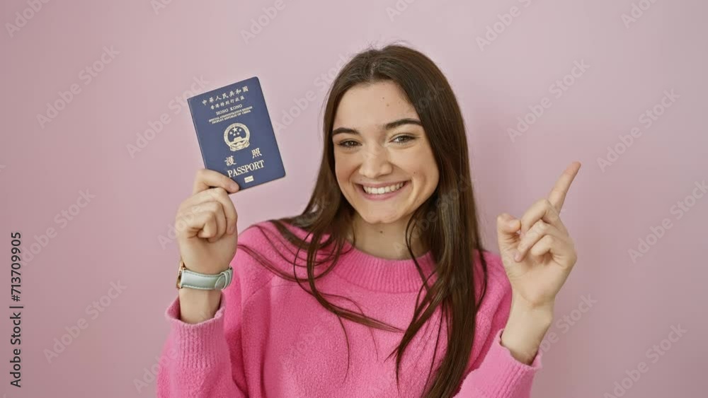 Joyful young hispanic woman, confidently showing off hong kong passport, gesturing to the side with a cheerful smile, isolated over a pink background. Joyful young hispanic woman, confidently showing off hong kong passport, gesturing to the side with a cheerful smile, isolated over a pink background.