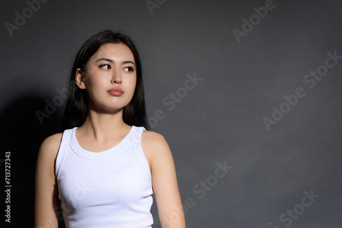 Brown-eyed Asian young girl model with clean smooth skin close-up posing on gray background, looking away. Beautiful young woman with dark hair in white T-shirt calmly watching