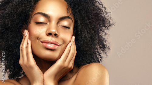 African American skincare model with a glowing healthy skin and curly hair is smiling and posing with a closed eyes against beige background. Beauty spa care treatment concept.