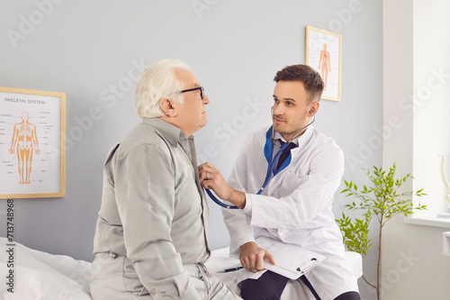 Male young doctor with stethoscope examining senior elderly man s lungs  breathing and heartbeat sitting on couch in exam room during medical checkup in clinic. Health care and medicine concept.