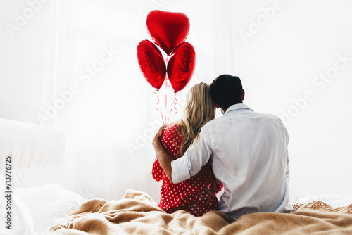 Couple. Love. Valentine's day. Back view of man and woman sitting on the bed, she is holding red heart-shaped balloons