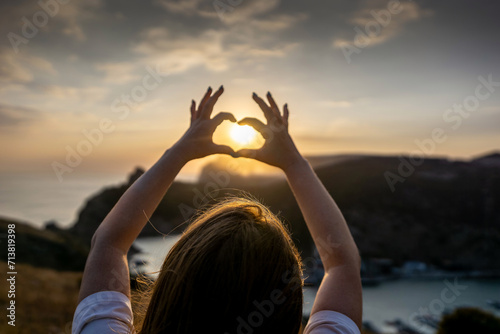 Happy woman on mountain peak  she makes heart shape with hands. Mountain  overlooking sea at sunset. Depicting love shape amidst scenic natural setting.