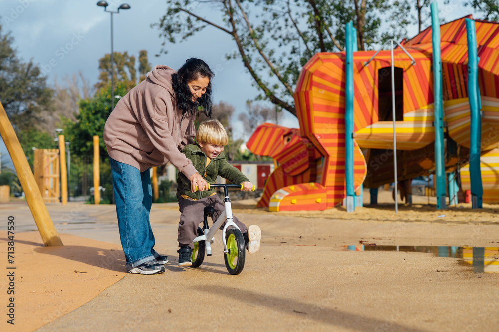 Obraz premium Mom teaches her little son to ride a bike without pedals. A boy on a balance bicycle. Along with the boy is the mother and supports the bike.