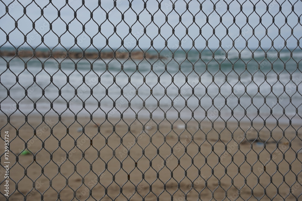 View of the winter beach through a mesh net fence. Mediterranean coast from behind metal wire fence. View of the winter beach through a mesh net fence. Mediterranean coast from behind metal wire fence.