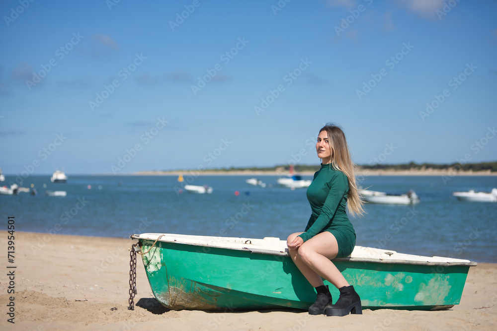 Young, pretty, blonde, green-eyed young woman in a green dress, sitting in a boat, looking at the camera, relaxed and calm, on the beach, with the sea in the background. Concept vacation, sun, travel