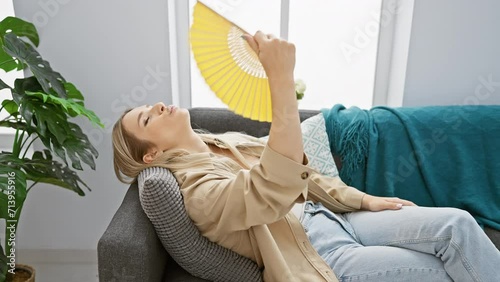 Stunning young blonde woman suffering in hot weather indoors, desperately fanning herself with a hand fan in her cosy home living room photo