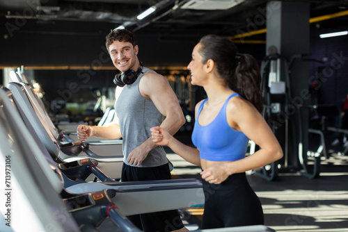 Excited young couple running on treadmills  looking at each other and smiling  enjoying cardio workout in modern gym interior  copy space
