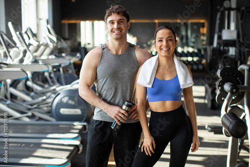 Happy man with water bottle and woman with towel on her shoulders posing after workout in gym and smiling, showing healthy lifestyle