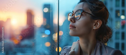 Thoughtful young businesswoman with glasses looking out office windows at city skyline. photo
