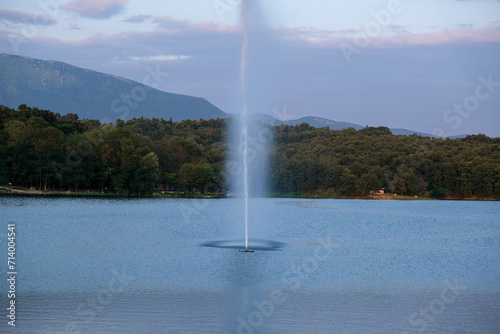 Tirana lake with a waterjet and breeze from a fountain, hills and green leafy trees on misty background, reflection on water surface, sunny spring day with abundant clouds in Tirana. High quality