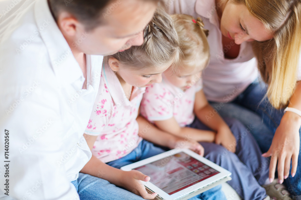 Happy family of mother, father and daughters sitting on a sofa at home having fun using a tablet computer Happy family of mother, father and daughters sitting on a sofa at home having fun using a tablet computer