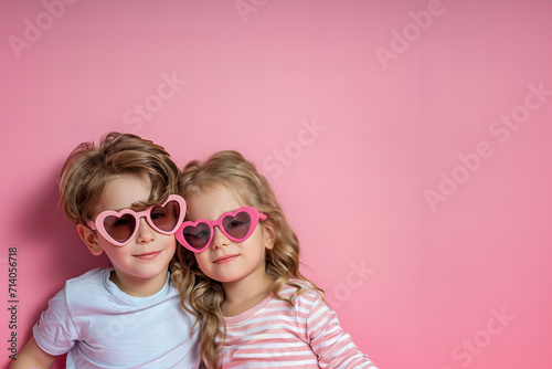 two cute kids, boy and girl wearing pink heart shaped sunglasses isolated on light pastel pink background with copy space © ALL YOU NEED studio