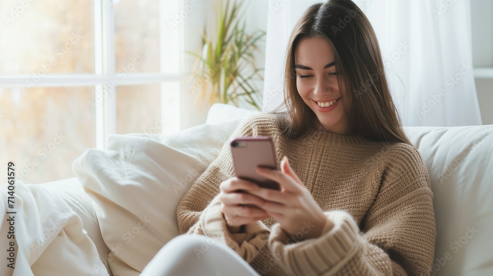 A happy woman in a light sweater is sitting on the couch, smiling, holding a phone in her hands