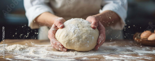 Hands kneading dough on a floured wood surface, flour dust in motion, baking process, fresh ingredients visible, culinary art, home cooking, chef in apron, pastry preparation, kitchen table. Banner. photo