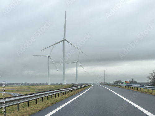 Tall green energy wind mills or turbines near a highway under a cloudy sky